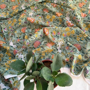 May include: A sage green fabric featuring a repeating floral pattern in shades of orange, yellow, and pink, with intricate vine and leaf details. The textile is draped in soft folds, showcasing the botanical print across the entire surface.
