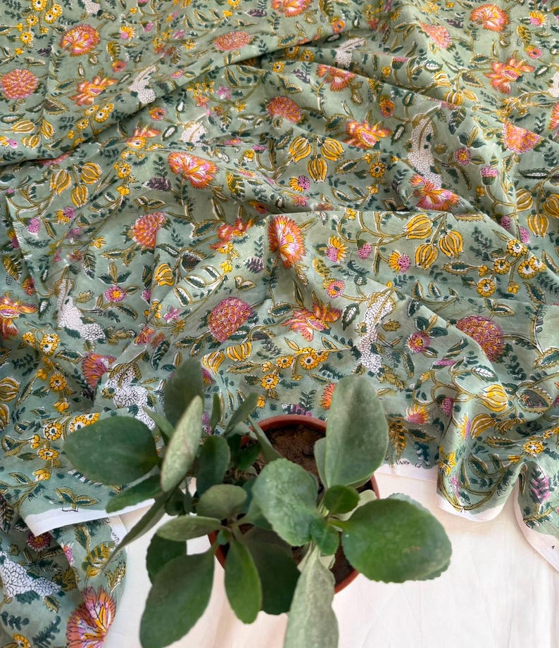 May include: A sage green fabric featuring a repeating floral pattern in shades of orange, yellow, and pink, with intricate vine and leaf details. The textile is draped in soft folds, showcasing the botanical print across the entire surface.