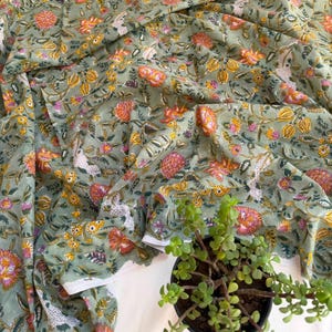 May include: A sage green fabric featuring a repeating floral pattern with orange, yellow, and pink blossoms, along with white and dark green leafy vines. The textile is draped in soft folds, displaying a dense, intricate botanical print throughout.