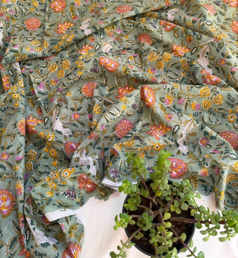 May include: A sage green fabric featuring a repeating floral pattern with orange, yellow, and pink blossoms, along with white and dark green leafy vines. The textile is draped in soft folds, displaying a dense, intricate botanical print throughout.