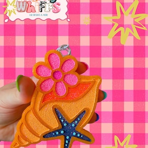 May include: An orange seashell-shaped car freshie featuring a textured surface, a bright pink flower accent at the top, and a dark blue starfish with light blue dots in the center. A silver metal ring is attached to the top for hanging purposes.