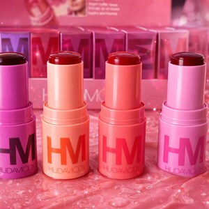 Puede incluir: Se muestran cuatro barras de labios de colores en tonos rosa y coral. Cada barra de labios tiene una tapa oscura y una base cilíndrica con las letras "HM" y el nombre de la marca "HUDAVOOD" impresos. El fondo es una superficie rosa y brillante.