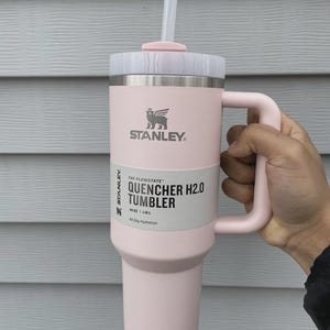 May include: A pink Stanley Quencher H2.0 tumbler with a handle and straw. The tumbler has a capacity of 40 oz (1.18 L) and features the Stanley logo. The text "All Day Hydration" is also visible.