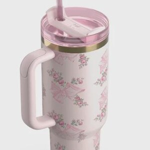 May include: A pink insulated travel mug with a handle and a straw. The mug has a floral and bow pattern in pink and white. The lid and handle are also pink, with a gold band at the top.