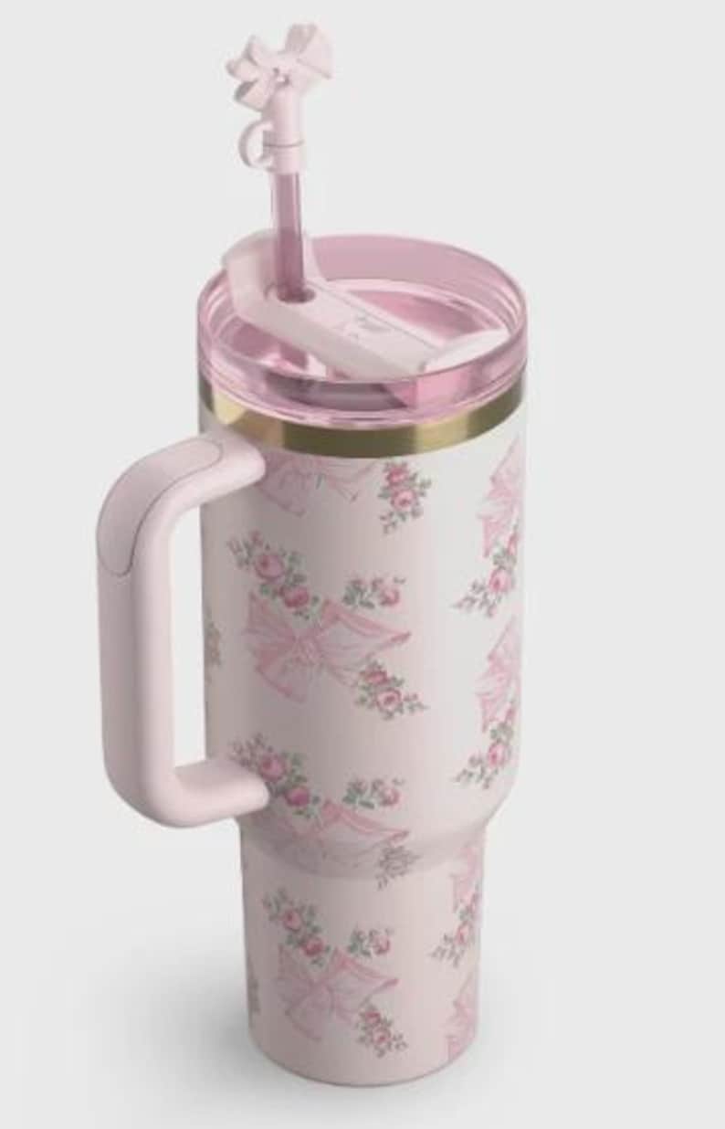 May include: A pink insulated travel mug with a handle and a straw. The mug has a floral and bow pattern in pink and white. The lid and handle are also pink, with a gold band at the top.