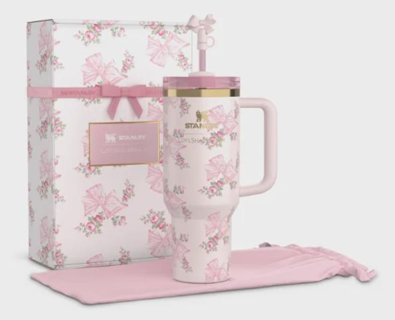 May include: A pink and white floral Stanley tumbler with a handle, straw, and lid, presented with a matching gift box and drawstring bag. The tumbler and box feature a repeating pattern of pink bows and roses. The gift box is tied with a pink ribbon.