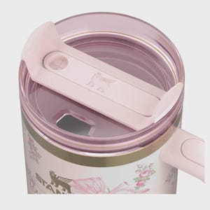 May include: A close-up of a pink and white floral Stanley tumbler. The tumbler has a pink lid and a pink sliding closure. The tumbler has a gold band around the middle and the Stanley logo is visible on the side.