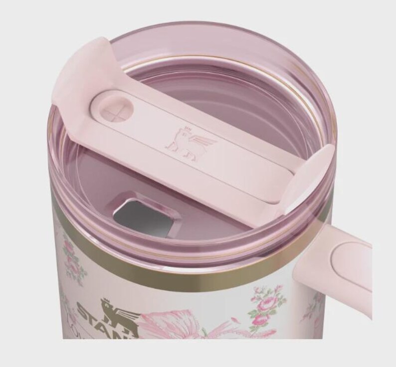 May include: A close-up of a pink and white floral Stanley tumbler. The tumbler has a pink lid and a pink sliding closure. The tumbler has a gold band around the middle and the Stanley logo is visible on the side.