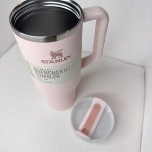 May include: A light pink Stanley Quencher H2.0 Flowstate tumbler with a handle and a lid. The tumbler has a silver rim and the Stanley logo. The lid is light pink and white. The tumbler is designed for all-day hydration.