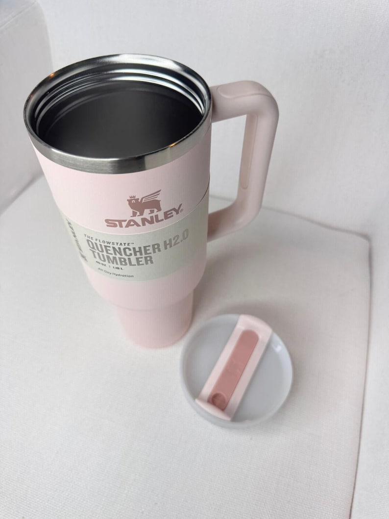 May include: A light pink Stanley Quencher H2.0 Flowstate tumbler with a handle and a lid. The tumbler has a silver rim and the Stanley logo. The lid is light pink and white. The tumbler is designed for all-day hydration.