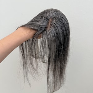 Könnte beinhalten: Ein Haarteil mit glattem, pfeffer-und-salz-grauem Haar und einem sichtbaren, hautähnlichen Scheitel. Das Haar ist gestuft und fällt in einem glatten, natürlichen Stil, wobei eine Mischung aus dunkelgrauen und silbernen Strähnen zu sehen ist.