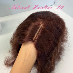 May include: A reddish-brown wig with a natural hairline fit. The wig has a center part and wavy texture. The text "Natural Hairline Fit" is written in pink cursive across the top of the image.