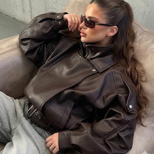 Puede incluir: Chaqueta de cuero marrón oscuro con cuello alto y cinturón. La chaqueta tiene cierre de cremallera y detalles de botones en los hombros. La persona lleva gafas de sol.