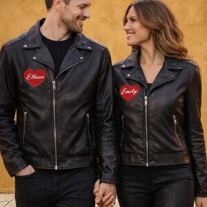 May include: Matching black leather jackets with silver zips and red heart patches. One jacket has "Ethan" embroidered on the heart, the other "Emily". The jackets are worn with black trousers. The couple is holding hands.