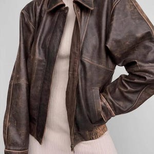 Peut inclure: Veste en cuir marron vieilli avec fermeture éclair, col et deux poches avant. La veste présente un aspect vintage et usé, avec des signes d'usure visibles. Veste de poids moyen, de style bombardier.
