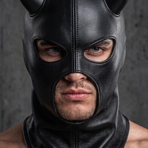 May include: A black leather hood featuring two prominent, curved horns extending from the top. The headgear includes a central vertical seam, a wide opening for the eyes, and a large circular cutout for the nose and mouth area, covering the head and neck.