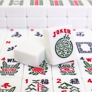 May include: A white Mahjong tile featuring the word JOKER in red text above a green circular graphic, positioned in front of another blank white tile. The tiles are arranged on a grid of additional Mahjong tiles with various red and green symbols.