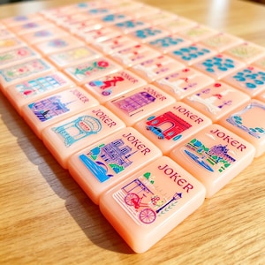 May include: A set of rectangular peach-colored Mahjong tiles arranged in rows. Each tile features a unique colorful illustration, such as Parisian landmarks, bicycles, and floral patterns, with the word JOKER printed in red above the graphic.