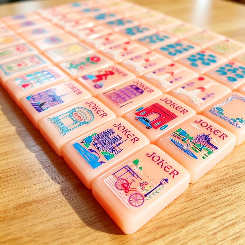 May include: A set of rectangular peach-colored Mahjong tiles arranged in rows. Each tile features a unique colorful illustration, such as Parisian landmarks, bicycles, and floral patterns, with the word JOKER printed in red above the graphic.