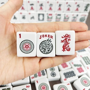 May include: Three white rectangular Mahjong tiles featuring black and red ink designs. The left tile displays the number 1 and a circular pattern, the center tile shows the word JOKER with a circular graphic, and the right tile depicts a red dragon.