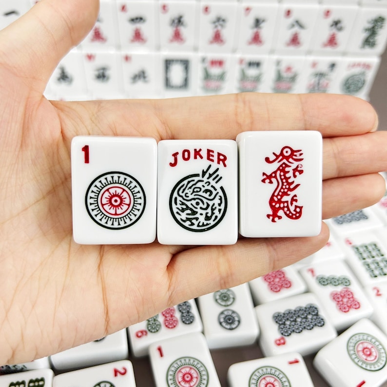 May include: Three white rectangular Mahjong tiles featuring black and red ink designs. The left tile displays the number 1 and a circular pattern, the center tile shows the word JOKER with a circular graphic, and the right tile depicts a red dragon.