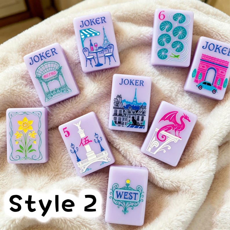 May include: A collection of nine rectangular light purple mahjong tiles featuring various colorful illustrations including a cafe, the Eiffel Tower, a dragon, a flower, and the text JOKER, 6, 5, and WEST. The tiles are arranged on a soft textured fabric.