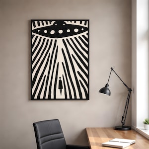Op de afbeelding: Een ingelijste wandkunstprint met een zwart-witte grafische illustratie. Het ontwerp toont een vliegende schotel die boven een klein, eenzaam figuur zweeft, met gedurfde verticale lijnen die naar beneden stralen om een gevoel van licht te creëren.