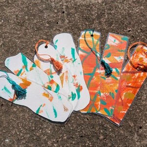 May include: A collection of handmade bookmarks with abstract designs. The bookmarks feature white, orange, and teal paint strokes. Each bookmark has a tassel attached with a string. The bookmarks are arranged on a textured, gray surface.