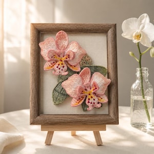 May include: A rectangular wooden frame containing a fabric panel with two embroidered pink and yellow orchid flowers and green leaves. The frame rests on a small wooden easel, displaying detailed thread work with textured petals and foliage.