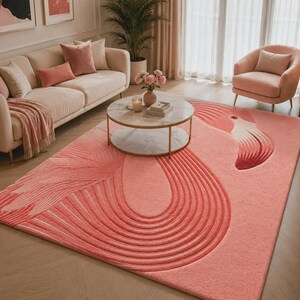 Puede incluir: Una alfombra rosa rectangular grande con un diseño de flamenco. La alfombra presenta una cabeza y cuello de flamenco con un patrón de plumas detallado en tonos de rosa y blanco. La alfombra está colocada en una sala de estar con un sofá, un sillón y una mesa de centro.
