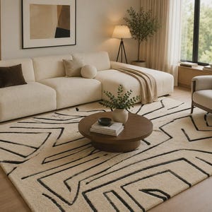 Puede incluir: Una alfombra rectangular con una base de color crema y un patrón geométrico de líneas negras audaces. El diseño consiste en formas angulares y líneas interconectadas que se extienden por toda la superficie de la alfombra texturizada.
