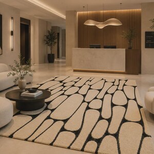 Puede incluir: Una alfombra rectangular grande con un fondo de color crema y un patr&oacute;n repetitivo de formas negras irregulares y redondeadas. La alfombra est&aacute; situada en un vest&iacute;bulo con paredes claras, muebles en tonos crema y una mesa circular oscura.