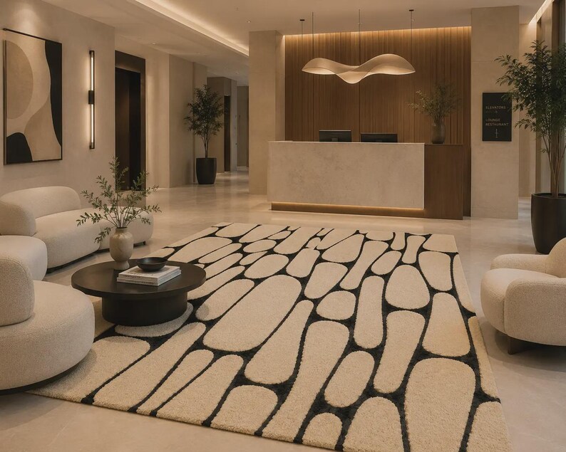 Puede incluir: Una alfombra rectangular grande con un fondo de color crema y un patr&oacute;n repetitivo de formas negras irregulares y redondeadas. La alfombra est&aacute; situada en un vest&iacute;bulo con paredes claras, muebles en tonos crema y una mesa circular oscura.