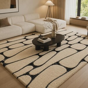 Puede incluir: Una alfombra rectangular con una superficie de pelo largo en color crema y un patr&oacute;n repetitivo de formas redondeadas similares a guijarros, delineadas en gris carb&oacute;n oscuro. La alfombra cubre una gran parte del suelo y tiene un acabado afelpado.