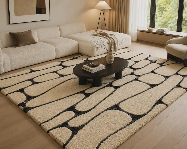 Puede incluir: Una alfombra rectangular con una superficie de pelo largo en color crema y un patr&oacute;n repetitivo de formas redondeadas similares a guijarros, delineadas en gris carb&oacute;n oscuro. La alfombra cubre una gran parte del suelo y tiene un acabado afelpado.