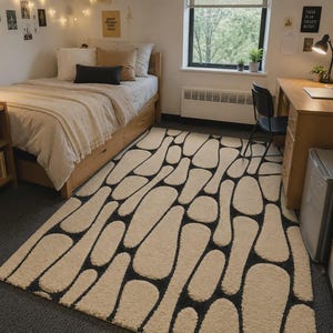 Puede incluir: Una alfombra rectangular con un patr&oacute;n repetitivo de formas redondeadas color crema, similares a guijarros, separadas por finas l&iacute;neas de color gris carb&oacute;n oscuro. La alfombra tiene una superficie texturizada y copetuda que cubre el suelo entre la cama y el escritorio.