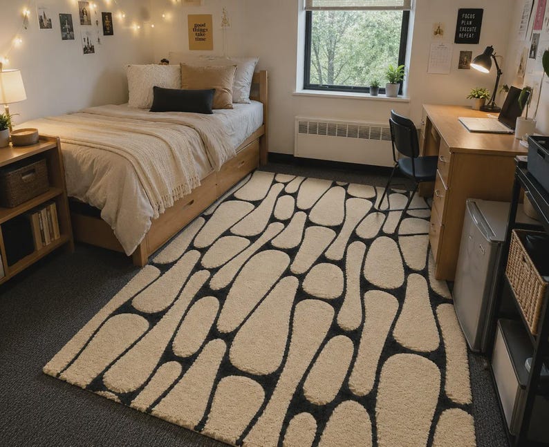 Puede incluir: Una alfombra rectangular con un patr&oacute;n repetitivo de formas redondeadas color crema, similares a guijarros, separadas por finas l&iacute;neas de color gris carb&oacute;n oscuro. La alfombra tiene una superficie texturizada y copetuda que cubre el suelo entre la cama y el escritorio.