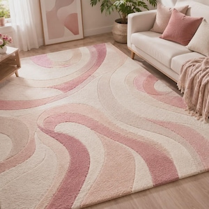 Puede incluir: Una alfombra rectangular grande con un diseño ondulado en tonos rosa y crema. La alfombra está colocada en un salón, con un sofá, cojines y objetos decorativos visibles en el fondo. El diseño de la alfombra añade un elemento suave y texturizado a la habitación.