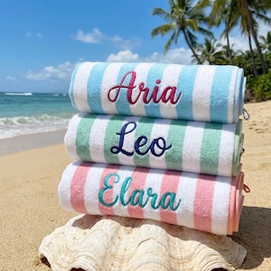 May include: Three rolled beach towels stacked vertically, each featuring white and colored stripes. The top towel is light blue with the name Aria embroidered in pink. The middle towel is mint green with the name Leo in navy. The bottom towel is pink with the name Elara in teal.