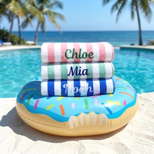 Könnte beinhalten: Drei gefaltete Strandtücher gestapelt auf einem blauen, donut-förmigen aufblasbaren Pool-Schwimmring. Das oberste Handtuch ist rosa-weiß gestreift mit dem Namen Chloe in Grün. Das mittlere ist grün-weiß gestreift mit Mia in Blau. Das untere ist blau-weiß gestreift mit Noah in Weiß.