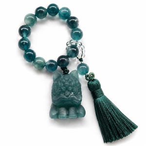 May include: A teal beaded bracelet with a carved cat pendant and a green tassel. The bracelet features large, round beads and a detailed cat figurine. The tassel is a deep green colour, adding a touch of elegance to the piece.
