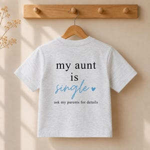 May include: A light gray heathered short-sleeve t-shirt featuring the black text "my aunt is", the blue script text "single" with a small blue heart, and the black text "ask my parents for details" printed on the front center of the garment.