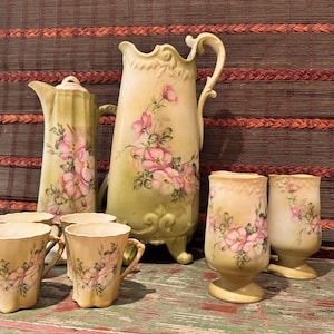 May include: A porcelain beverage set featuring a tall pitcher, a lidded carafe, and four matching cups. Each piece is finished in a pale yellow glaze with hand-painted pink floral motifs, scalloped edges, and ornate, curved handles on the vessels.