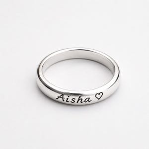 Peut inclure: Une bague anneau en métal argenté poli portant le prénom Aisha gravé en écriture cursive, suivi d'un petit symbole de cœur. La bague présente une surface lisse et arrondie avec une finition métallique uniforme sur tout son design circulaire.