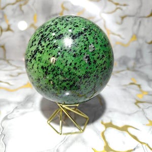Könnte beinhalten: Eine grüne Kugel mit schwarzen Sprenkeln, präsentiert auf einem goldenen geometrischen Ständer. Der polierte Stein hat einen leuchtenden Grünton mit dunklen Flecken. Der Ständer besteht aus dünnem, goldfarbenem Metall.