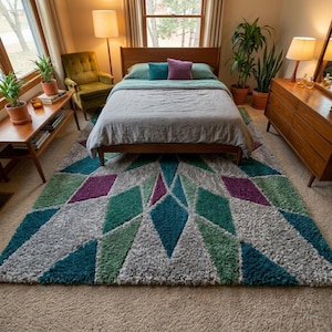 May include: A bedroom scene featuring a mid-century modern aesthetic. A large, geometric patterned rug in shades of gray, teal, green, and purple dominates the floor. A wooden bed frame, dresser, and side table complement the rug. Green plants and a green armchair add pops of color.