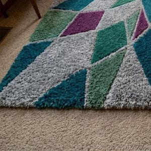 May include: A colorful geometric area rug with a shaggy texture. The rug features diamond shapes in teal, purple, green, and gray. The rug is placed on a beige carpeted floor.