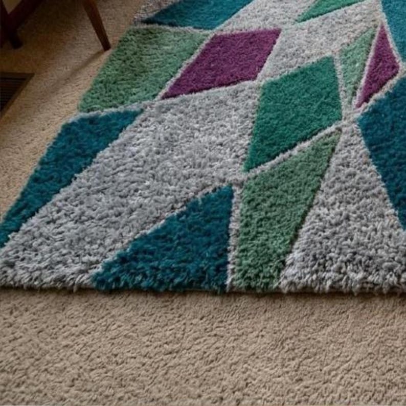 May include: A colorful geometric area rug with a shaggy texture. The rug features diamond shapes in teal, purple, green, and gray. The rug is placed on a beige carpeted floor.