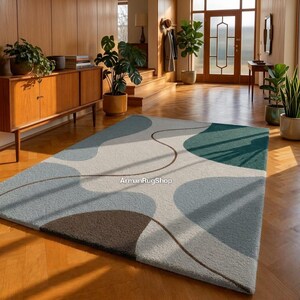 May include: A rectangular area rug with an abstract design in shades of blue, green, and brown. The rug features a light beige background with organic shapes and a curved brown line. The rug is placed on a wooden floor.