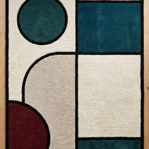 May include: A rectangular area rug featuring a geometric pattern with bold black outlines. The design includes a teal circle, a deep red semicircle, and large blocks of cream, beige, and dark teal. The surface has a soft, textured pile finish.